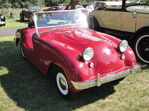 Deep red doorless roadster with a fold-flat windscreen and round headlamps, parked on a lawn. Opens a larger view