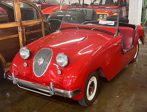 Red doorless roadster with an oval grille and round headlamps on the nose, a wood-bodied car beside it. Opens a larger view