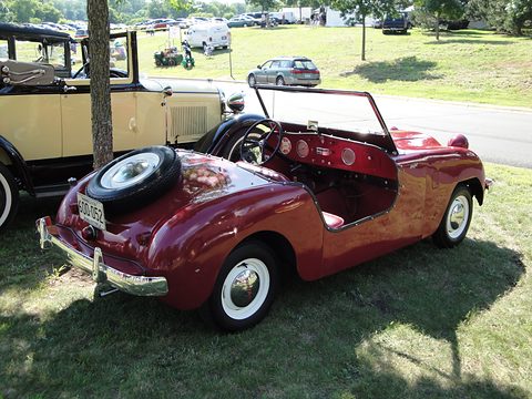 Deep red roadster from the left rear with its spare wheel on the tail and a dashboard in body colour. Opens a larger view
