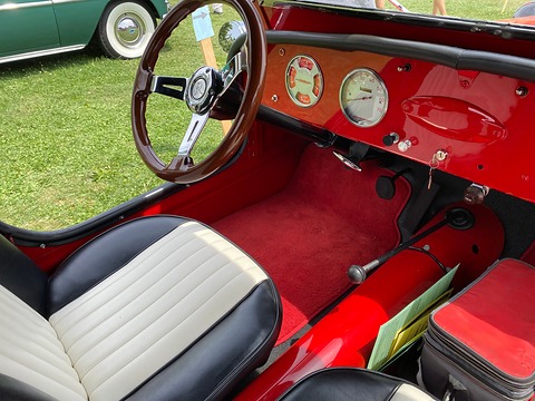 Red painted dashboard with two round dials, a wood-rimmed wheel, red carpet and a pleated white seat. Opens a larger view