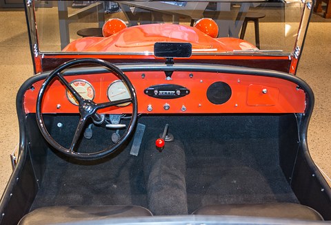 Orange dashboard with two dials and a large black steering wheel above a dark floor and seats. Opens a larger view