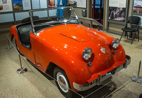Orange doorless roadster with round headlamps and a small crest, roped off on a terrazzo floor. Opens a larger view