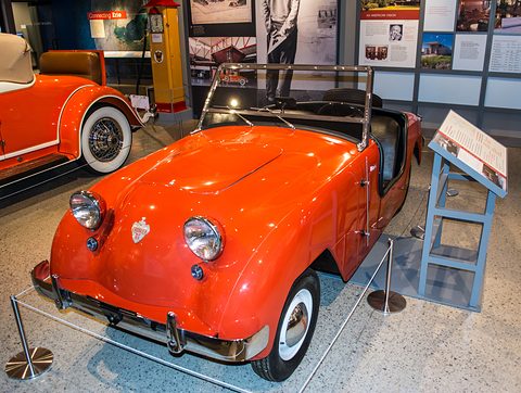 Orange doorless roadster with its windscreen up, display boards and an orange car with whitewalls behind. Opens a larger view