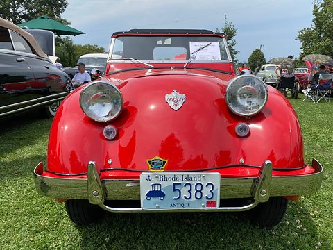 Front of a red roadster: two large round headlamps, a small crest and a Rhode Island plate. Opens a larger view