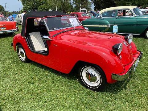 Red doorless roadster with a black soft top and white seats, parked on grass among other cars. Opens a larger view