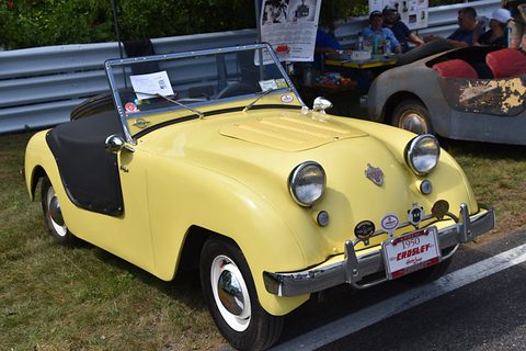 Pale yellow doorless roadster with a black interior and twin headlamps, parked by a crash barrier. Opens a larger view