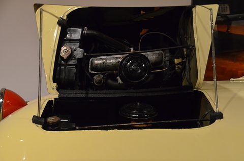 Engine bay under a raised cream bonnet, a small four-cylinder engine sitting behind the front axle. Opens a larger view