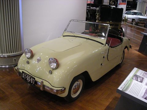 Cream doorless roadster with a red interior on a wooden museum floor, a display panel beside it. Opens a larger view