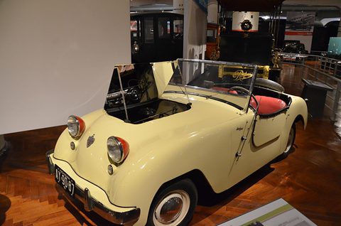 Cream doorless roadster with its bonnet open and a red interior, on a parquet floor among exhibits. Opens a larger view