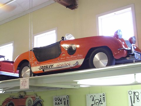 Orange roadster lettered CROSLEY Hotshot with a black tonneau, high on a storage shelf above tools. Opens a larger view