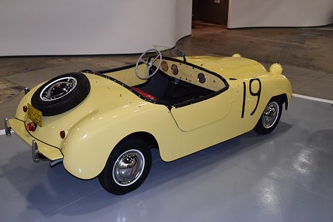 Cream doorless roadster with a black number 19 on its side and a spare wheel on the tail, on a polished floor. Opens a larger view