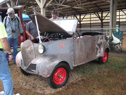 Grey bare-metal small open car with red wheels and its bonnet raised, on grass, men looking at it. Opens a larger view