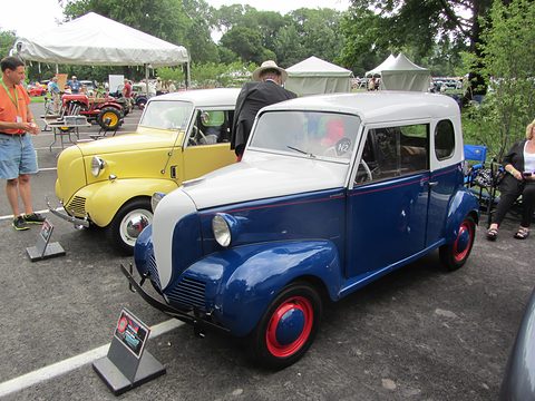 Small blue two-door with a white top and red wheels on tarmac, a yellow Crosley and a white marquee behind. Opens a larger view