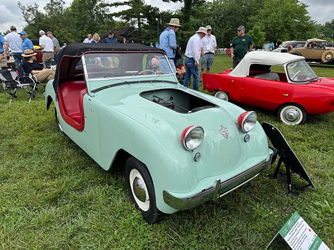 Mint-green roadster with a red interior parked on a lawn, visitors and a red car behind. Opens a larger view