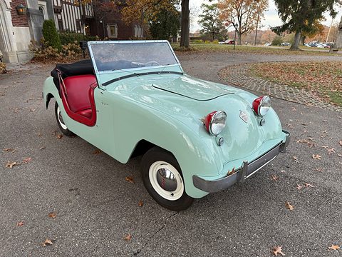 Mint-green roadster with a red interior and black top folded, on a gravel drive before a half-timbered house. Opens a larger view