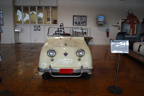 Cream roadster on a dark wooden floor in a large hall, a chequered flag on its windscreen. Opens a larger view