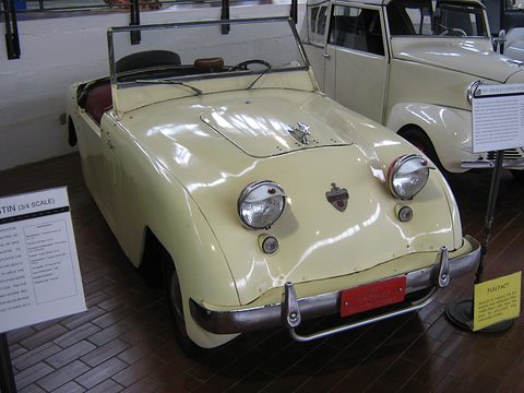Cream roadster with twin round headlamps and a small crest on the nose, a white van and display board beside it. Opens a larger view