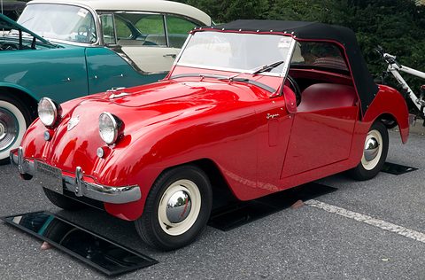 Red roadster with a black soft top and a red interior, parked on tarmac beside a turquoise car. Opens a larger view