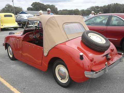 Red roadster from the left rear with a tan soft top, a door and a spare wheel on the tail. Opens a larger view