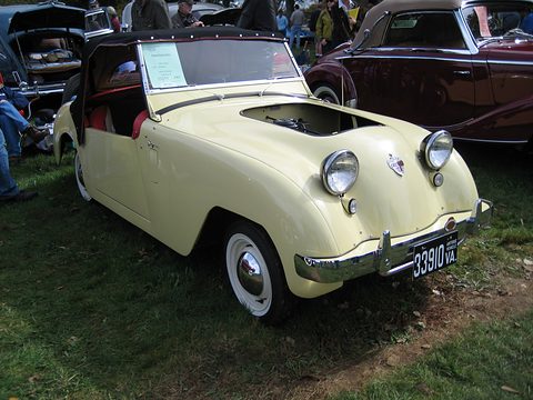 Cream roadster with a red interior, a black top and a card on its windscreen, parked on grass among show cars. Opens a larger view