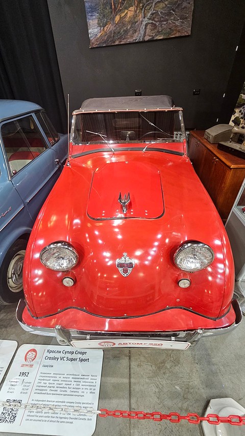 Red roadster from the front with twin headlamps and a crest, an information panel on the floor in front. Opens a larger view