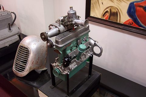 Small four-cylinder engine with a supercharger and twin carburettors on a black stand beside a grille cowl. Opens a larger view