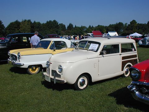 White small estate parked on grass beside a two-tone Nash Metropolitan, a crowd and trees behind. Opens a larger view