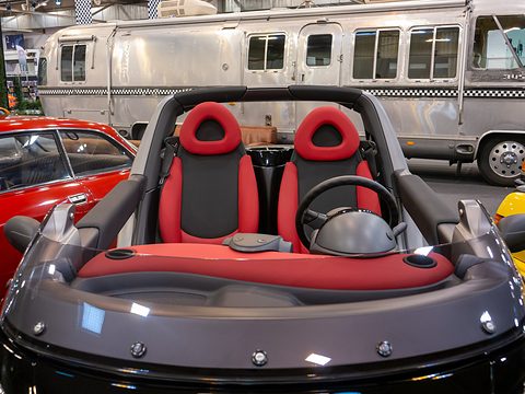 Two red and black bucket seats and a steering wheel behind a low screen, an aluminium motorhome in the hall behind. Opens a larger view