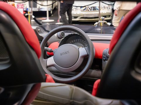 View between two red seats onto a grey steering wheel with a Smart badge, round instruments and the low screen. Opens a larger view