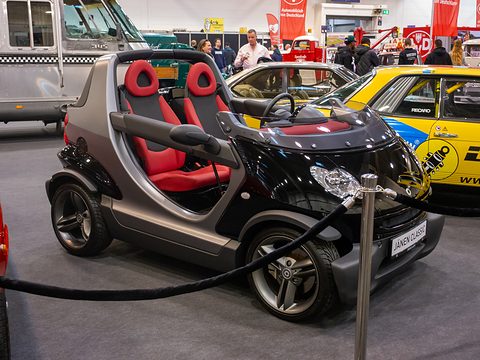 Black and grey doorless two-seater with red seats behind a rope barrier, among other cars and visitors in an exhibition hall. Opens a larger view
