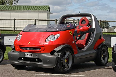 Doorless two-seater with a bright red front, prancing-horse badge and grey sides, parked by a fence on a grey day. Opens a larger view