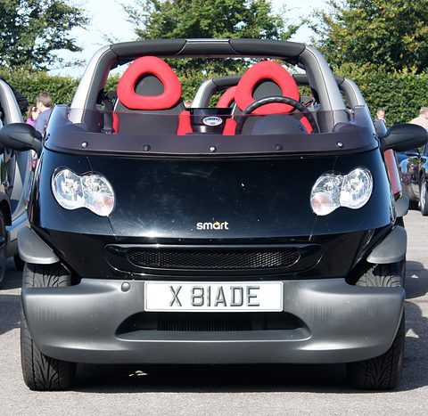 Head-on view of a black and silver doorless two-seater with red seats, twin headlamps and a silver grille. Opens a larger view