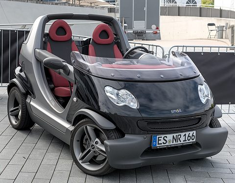 Grey and black doorless two-seater with red seats parked on paving in front of metal barriers.