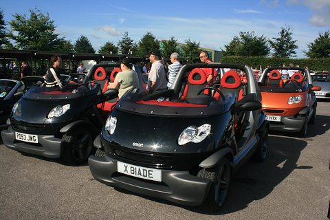 Several doorless two-seaters in black, orange and silver with red seats, parked side by side as visitors walk among them. Opens a larger view