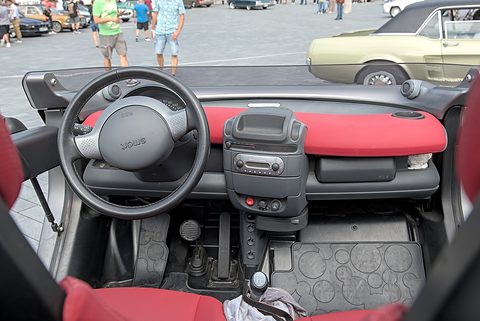 Looking down into an open two-seat cabin with red and black seats, a grey steering wheel and a patterned rubber floor.