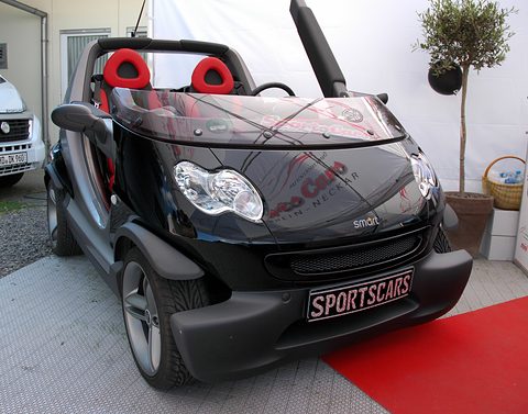 Black doorless two-seater with red seats on a red carpet, a dealer plate on its bumper, in a white tent beside a potted tree. Opens a larger view