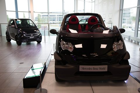 Black doorless two-seater with red seats on a display floor, a Mercedes-Benz World plaque in front and a small Smart city car behind. Opens a larger view