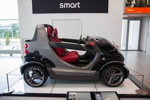 Black and grey doorless two-seater with red seats seen side-on on a white plinth behind a glass barrier, a Smart sign above. Opens a larger view