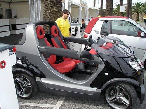 Grey and black doorless two-seater with red seats beside a white Smart city car, a man standing behind it. Opens a larger view