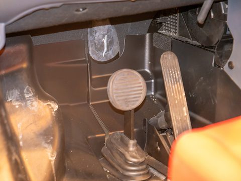 Footwell of a small car with two pedals, a gear-lever gaiter and a ribbed black rubber floor, lit warmly. Opens a larger view
