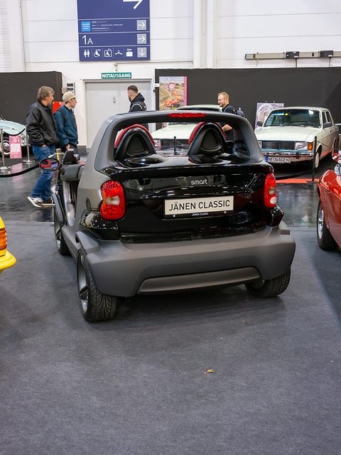 Rear of a black and grey doorless two-seater with round tail lamps and a dealer plate, on a carpeted stand among other classic cars. Opens a larger view