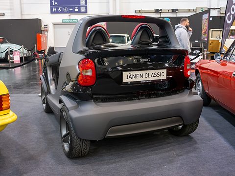 Black and grey doorless two-seater with red seats seen from behind and to one side, a yellow car and a red car beside it. Opens a larger view