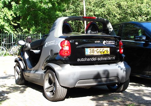 Rear of a black and silver doorless two-seater with a dealer web address across its tail, parked on paving under trees. Opens a larger view