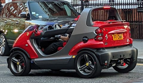 Red and grey doorless two-seater with a raised rear spoiler, driven past a large dark car and a brick building. Opens a larger view