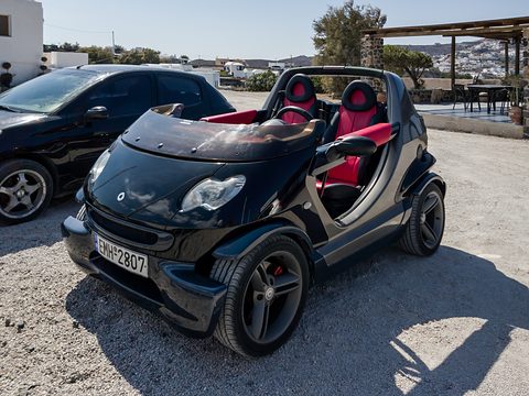 Black doorless two-seater with red seats parked on gravel in strong sun, white island houses on the hillside behind. Opens a larger view