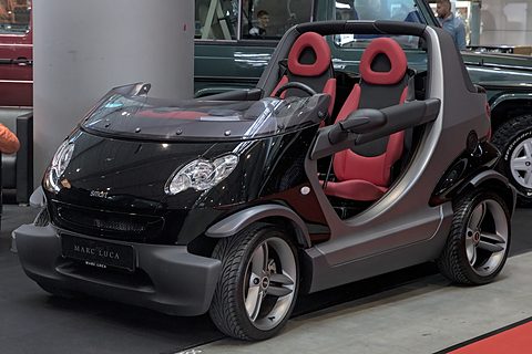 Black doorless two-seater with red seats and roll hoops on a show floor, other classic cars behind. Opens a larger view
