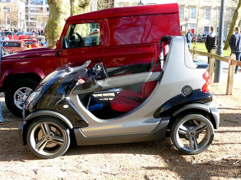 Silver and black doorless two-seater with red seats seen from the side, parked on gravel in front of a red Land Rover. Opens a larger view
