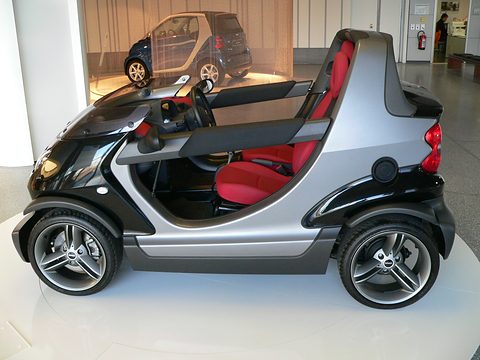 Silver and black doorless two-seater with red seats in profile on a round white plinth, a Smart city car behind a screen.