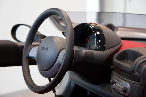 Close view of a grey leather steering wheel with a Smart badge, a black instrument pod and the centre console. Opens a larger view