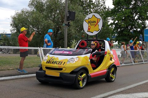 Yellow and red doorless two-seater with roll hoops and a round sign on a pole, driven past spectators behind metal barriers. Opens a larger view